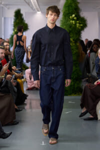 Male model walks the runway in a navy button-down shirt, wide dark jeans, and tan sandals, with a green plant backdrop and seated audience on both sides.