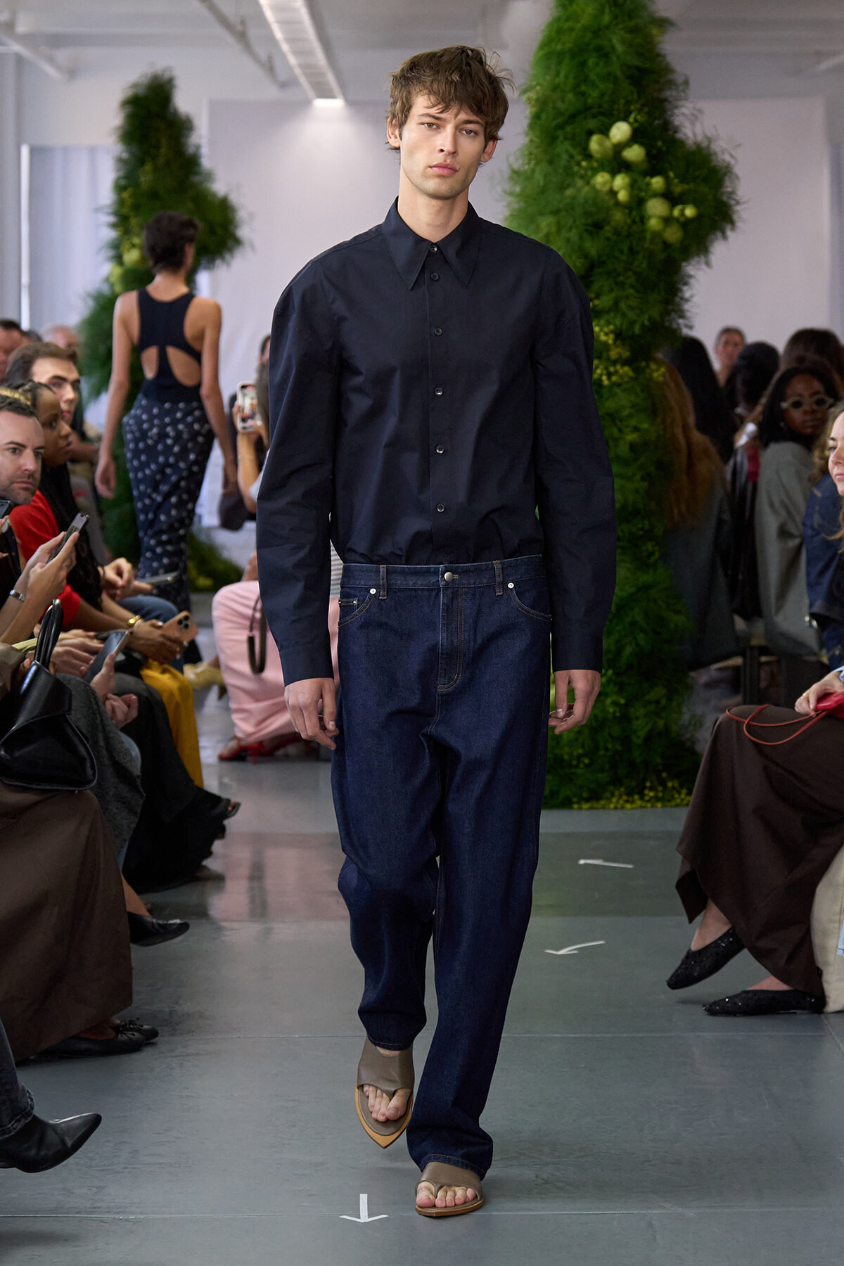 Male model walks the runway in a navy button-down shirt, wide dark jeans, and tan sandals, with a green plant backdrop and seated audience on both sides.