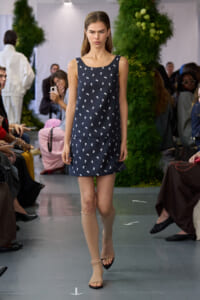 Model walks a fashion runway wearing a navy sleeveless dress with tiny white floral prints, paired with beige knee-high socks and sandals, audience in background.