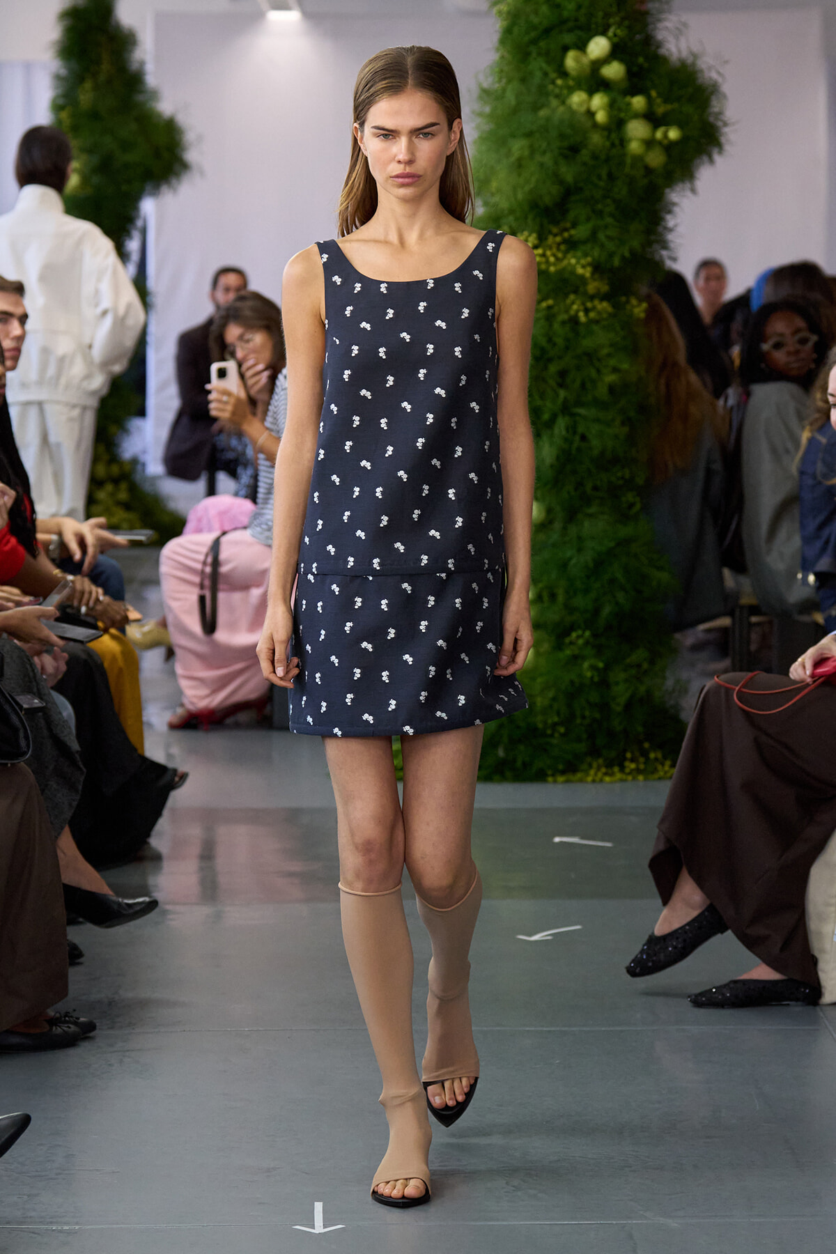 Model walks a fashion runway wearing a navy sleeveless dress with tiny white floral prints, paired with beige knee-high socks and sandals, audience in background.