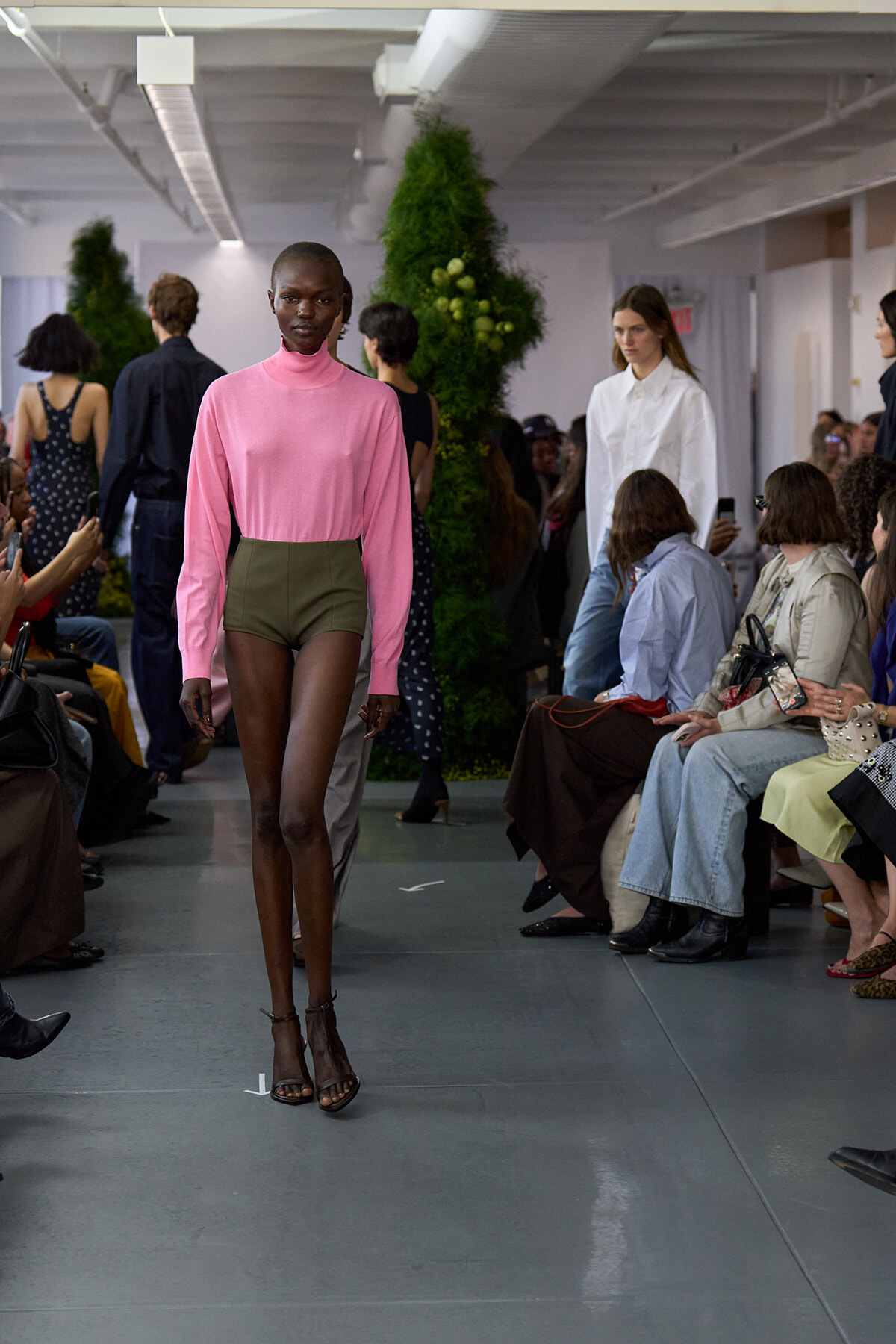 Model walks a fashion show runway in a pink turtleneck and olive-green high-waisted shorts; audience sits along the sides watching.
