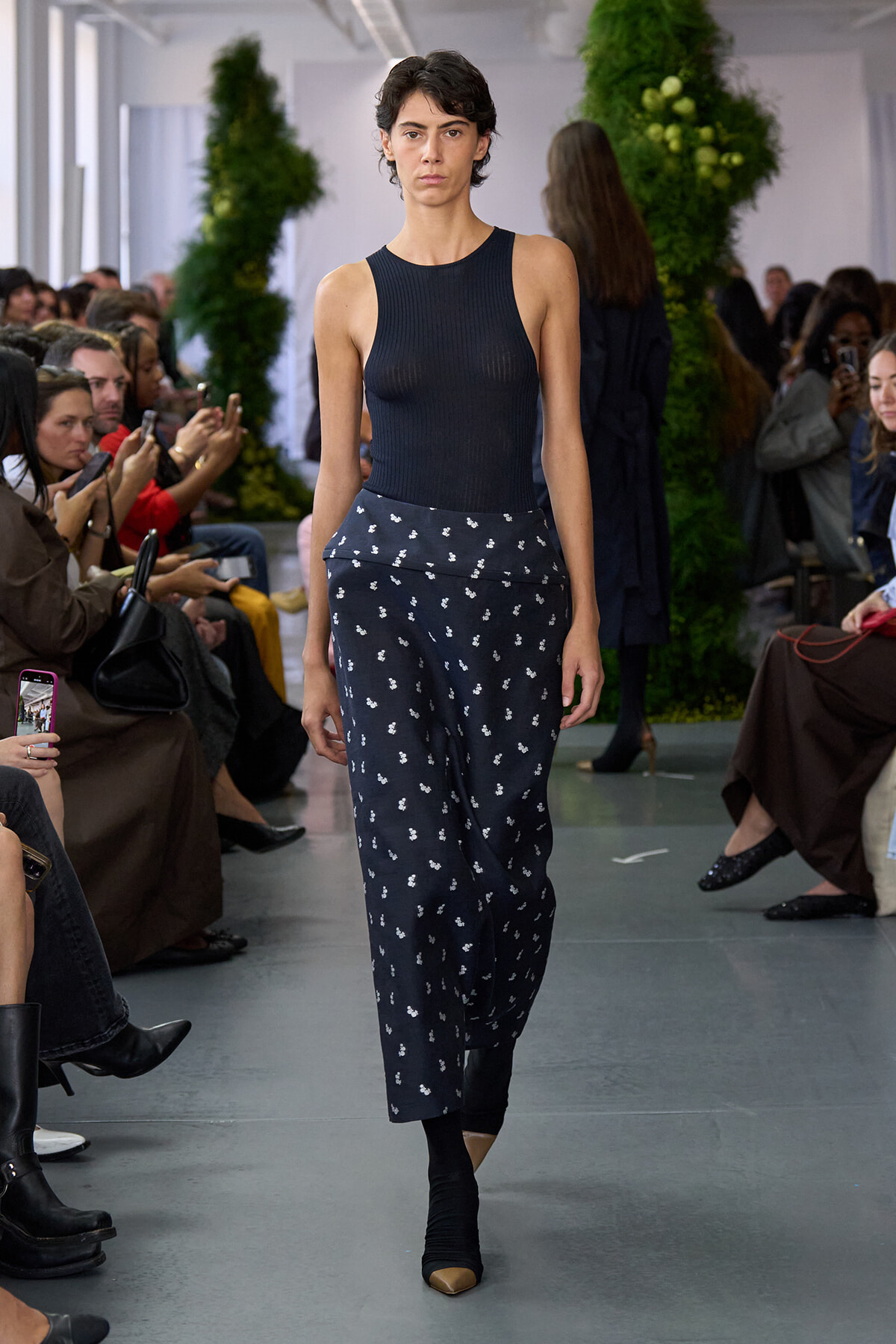 Model walks the runway in a navy sleeveless knit top and navy floral-print pants as seated audiences watch.