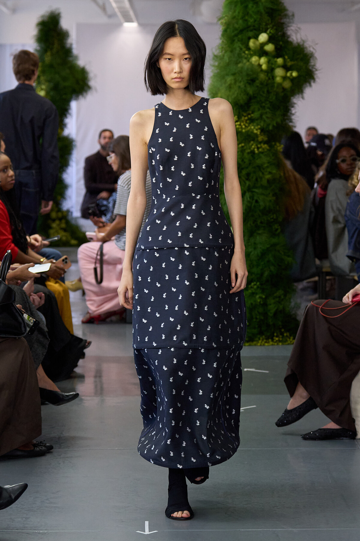 Model walking a navy sleeveless maxi dress with small white floral print on a runway, audience watching in the background.