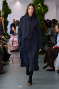 Female model walking the runway in a navy oversized coat with a high collar, paired with black tights and brown pointed-toe flats, audience and greenery in the background