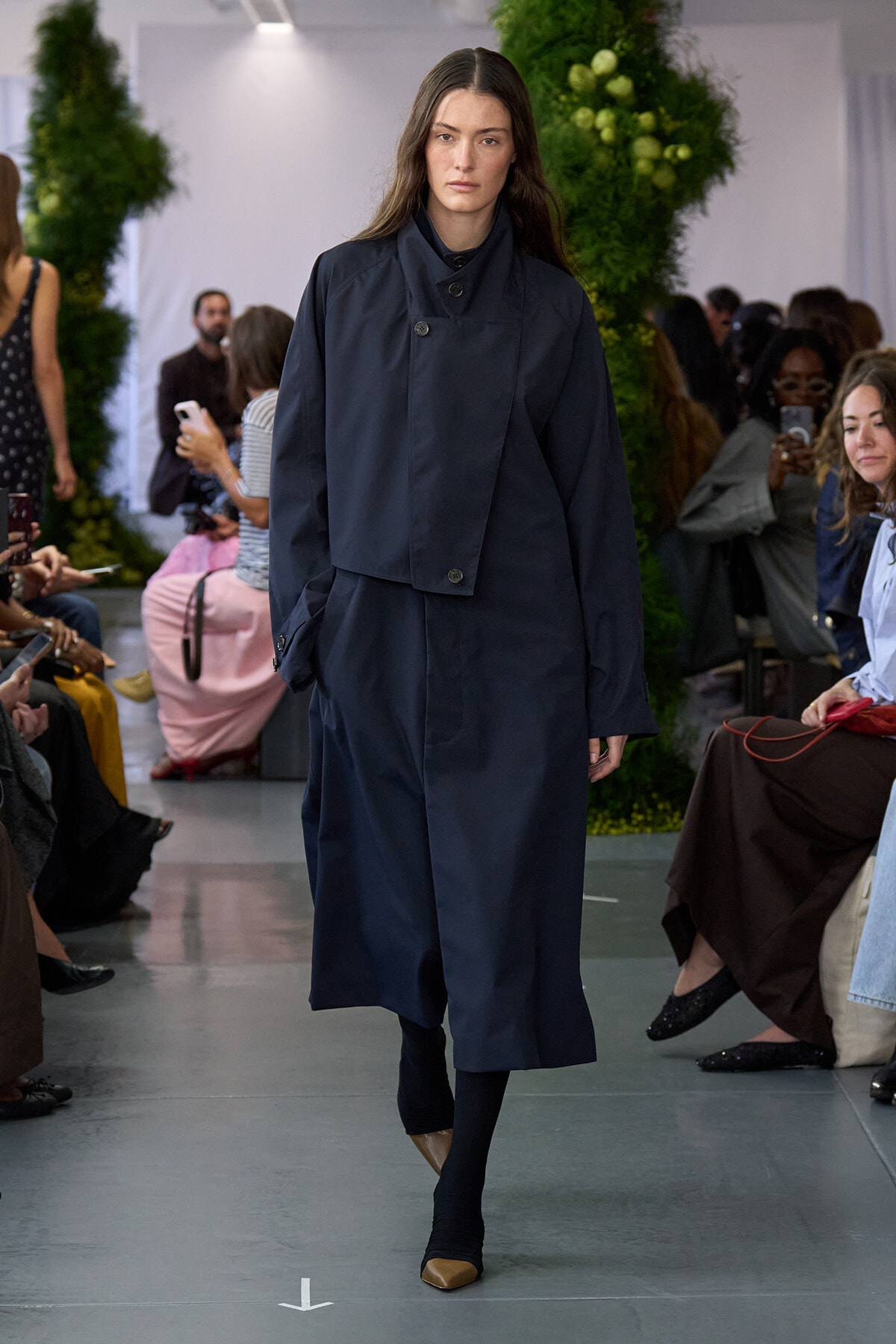 Female model walking the runway in a navy oversized coat with a high collar, paired with black tights and brown pointed-toe flats, audience and greenery in the background