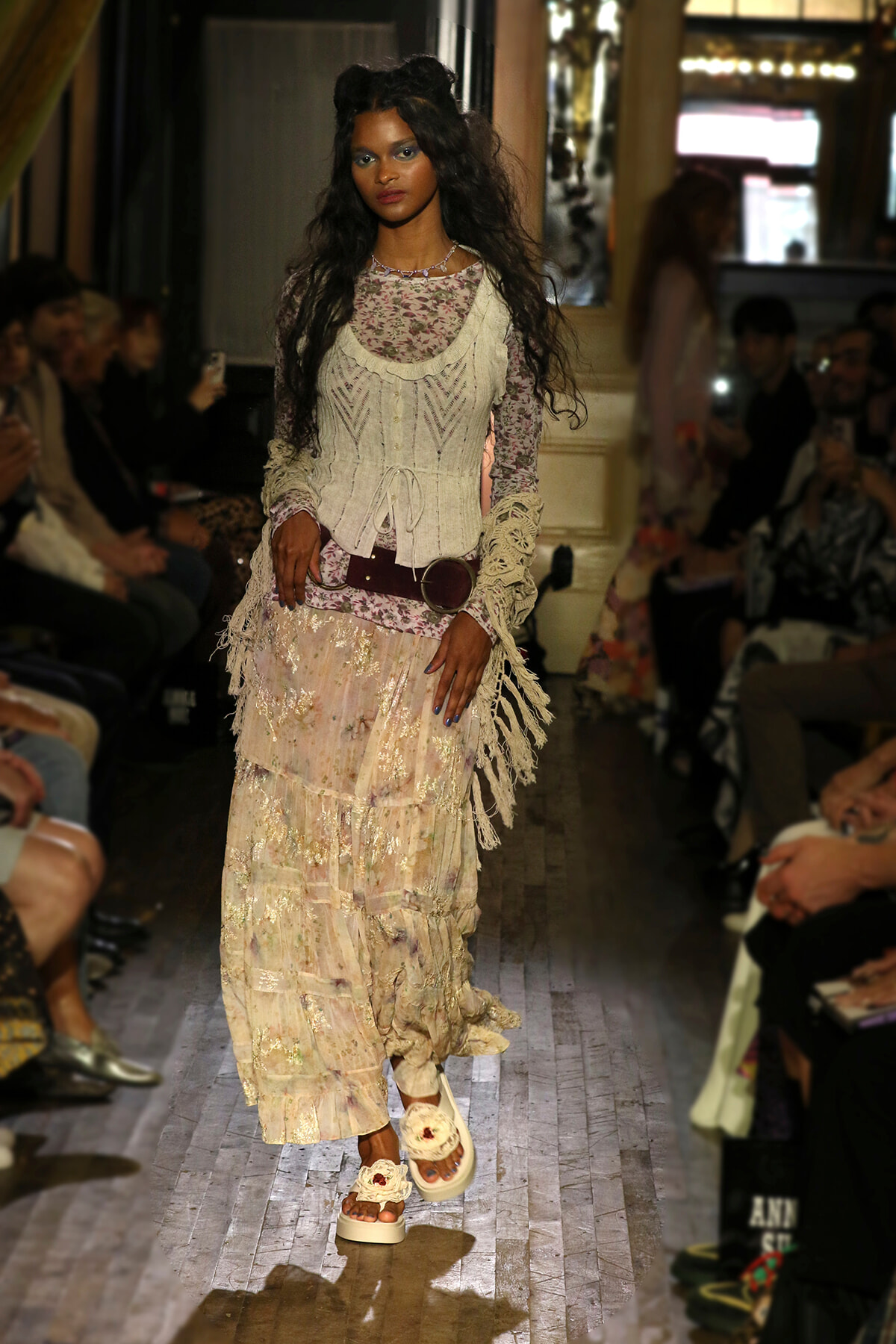 Model walks a boho-inspired runway wearing a cream lace layered dress with fringe, belt, and floral platform sandals.