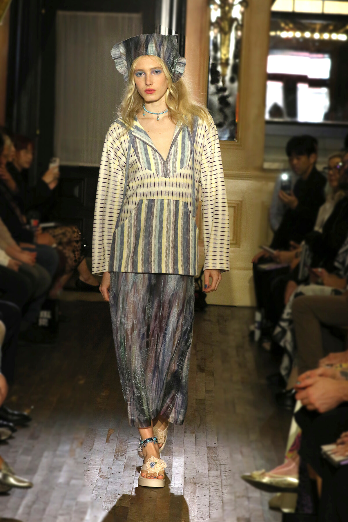 Model walks the runway in a striped, cream-and-blue top paired with a metallic, pleated skirt and matching headpiece.