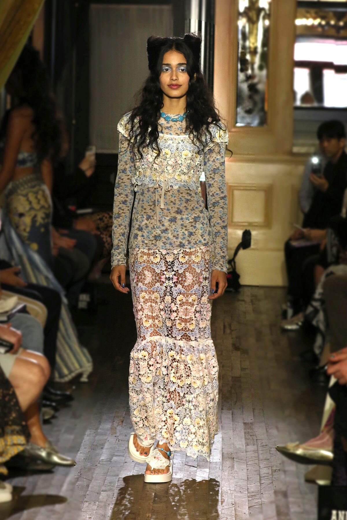 Runway model walks the catwalk in a pale blue floral lace dress with long sleeves and a turquoise beaded necklace.