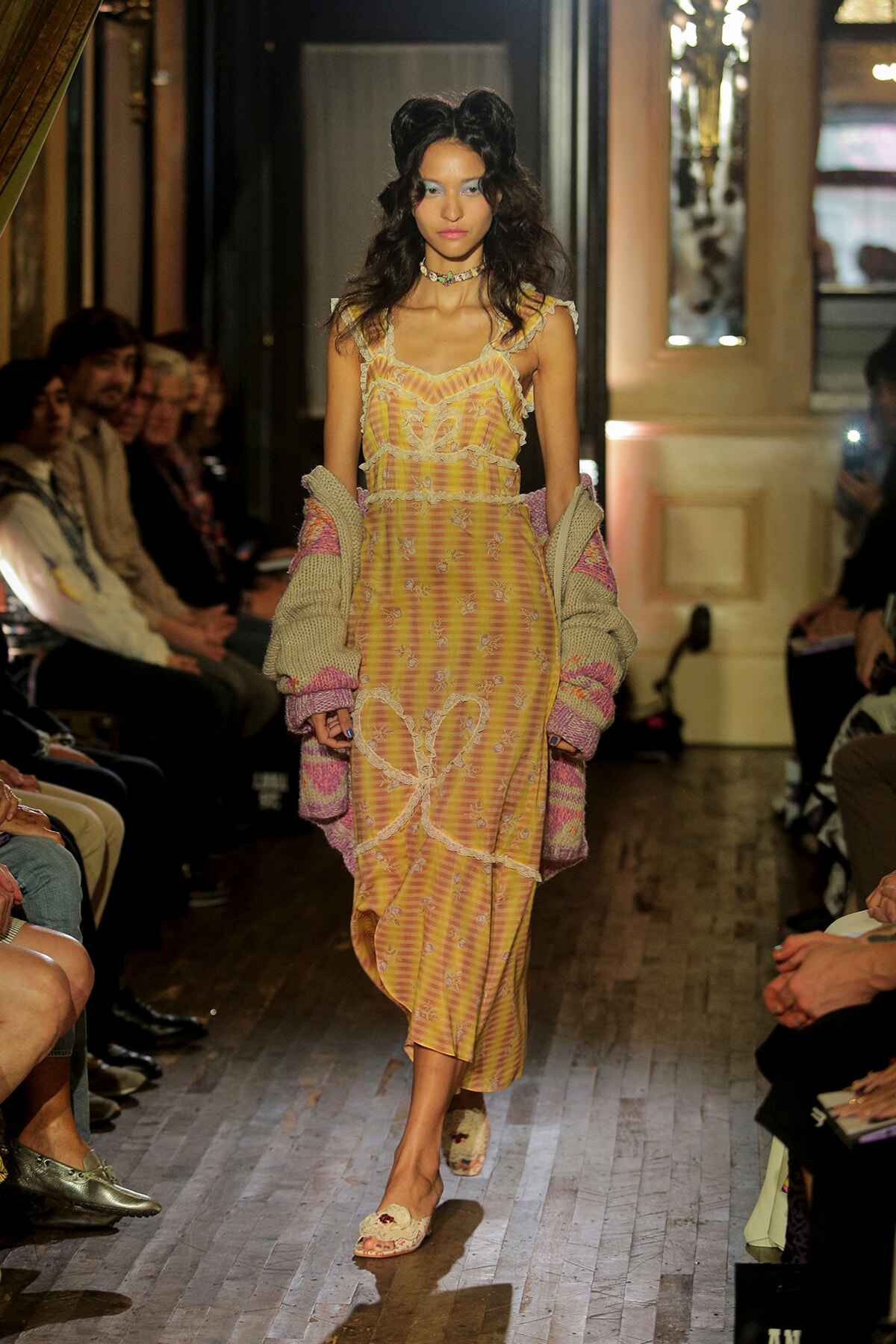 Model walking a fashion runway in a pastel yellow striped dress with lace trim and a chunky knit cardigan draped over her arms; audience seated along the runway watch.