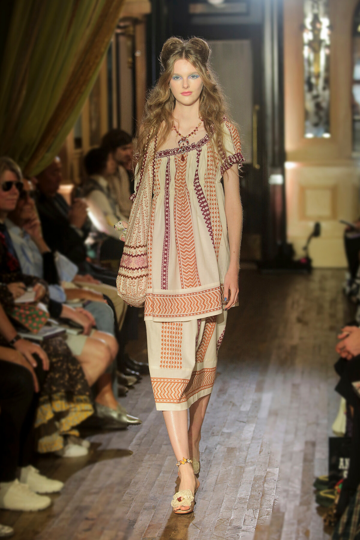 Model walks the runway in a cream bohemian dress with orange and burgundy geometric patterns, carrying a crocheted bag in a fashion-show setting.