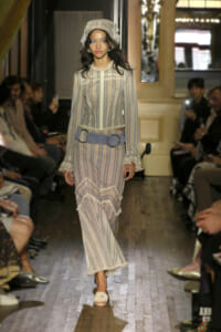 Model on a runway wearing a pale striped vintage dress with lace trim, a wide blue belt, and a headscarf.
