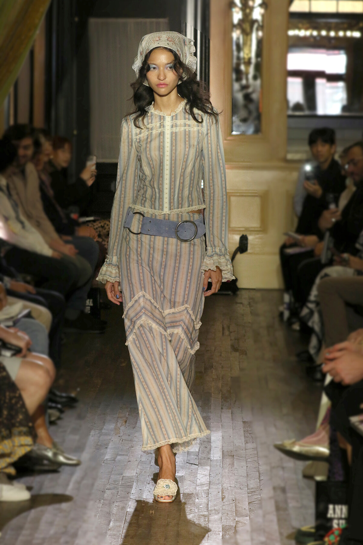 Model on a runway wearing a pale striped vintage dress with lace trim, a wide blue belt, and a headscarf.