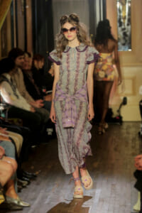 Model walking the runway in a lavender lace dress with purple accents, a tied waist wrap, and oversized sunglasses.