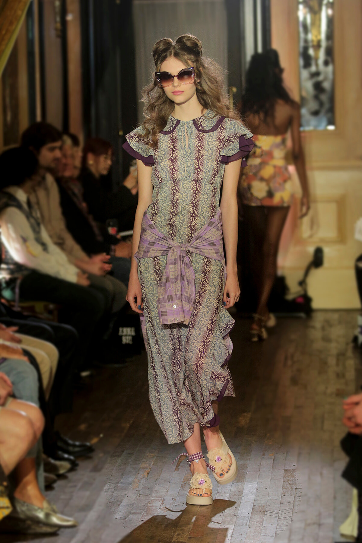 Model walking the runway in a lavender lace dress with purple accents, a tied waist wrap, and oversized sunglasses.