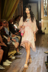 Model walking the runway in a pale pink lace dress, carrying a pink plush bag, with audience seated beside the catwalk.