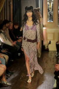 Model walks a floral-print long-sleeve dress with a wide burgundy belt on a runway lined with seated audience.