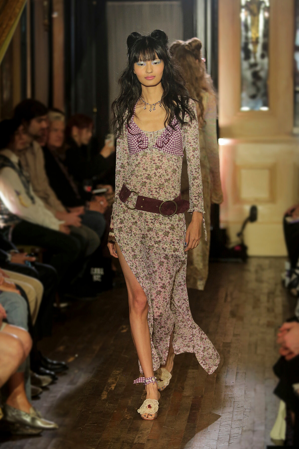 Model walks a floral-print long-sleeve dress with a wide burgundy belt on a runway lined with seated audience.
