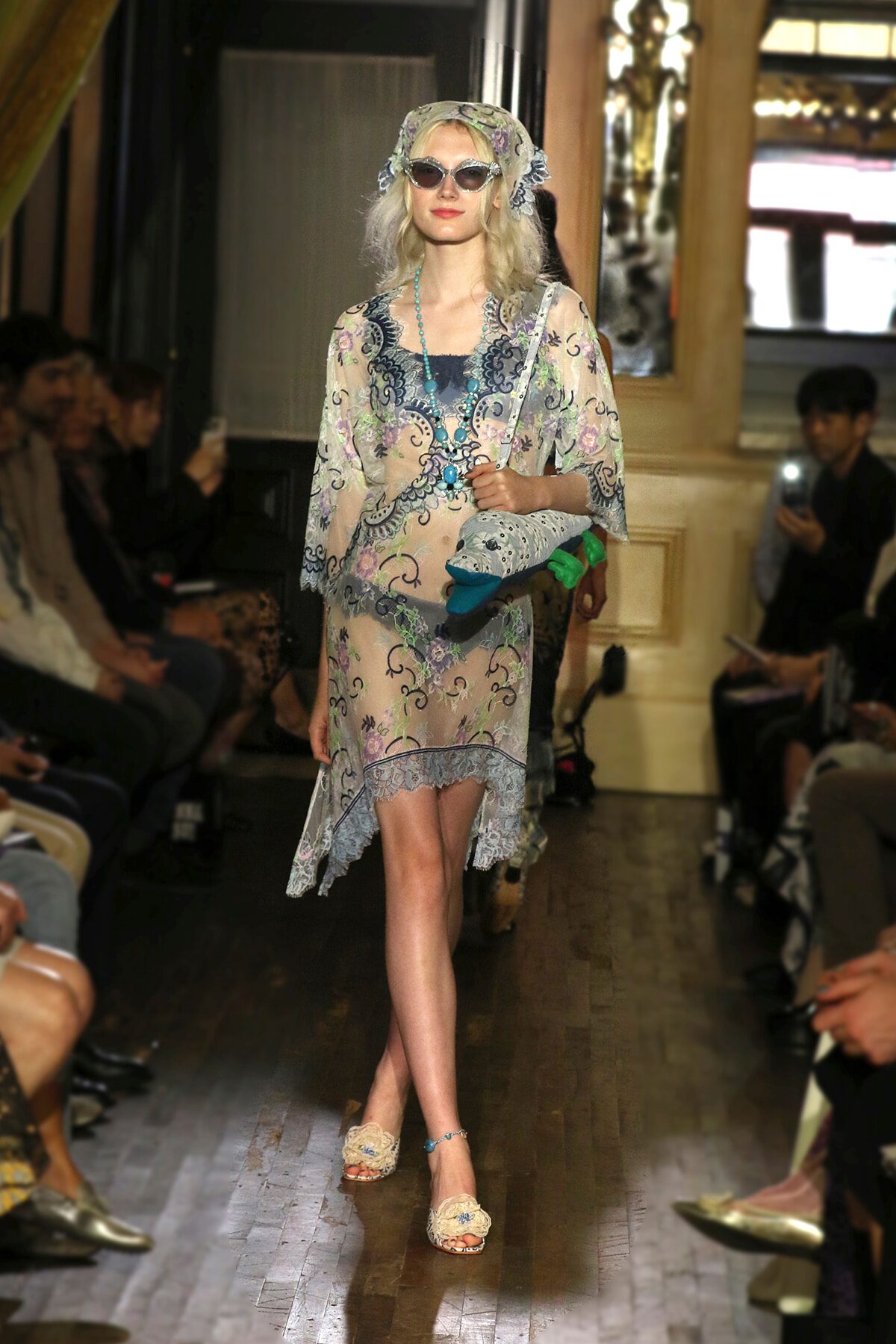 Model walking a runway in a sheer pastel floral lace dress with a headscarf, oversized sunglasses, and turquoise beaded necklace.