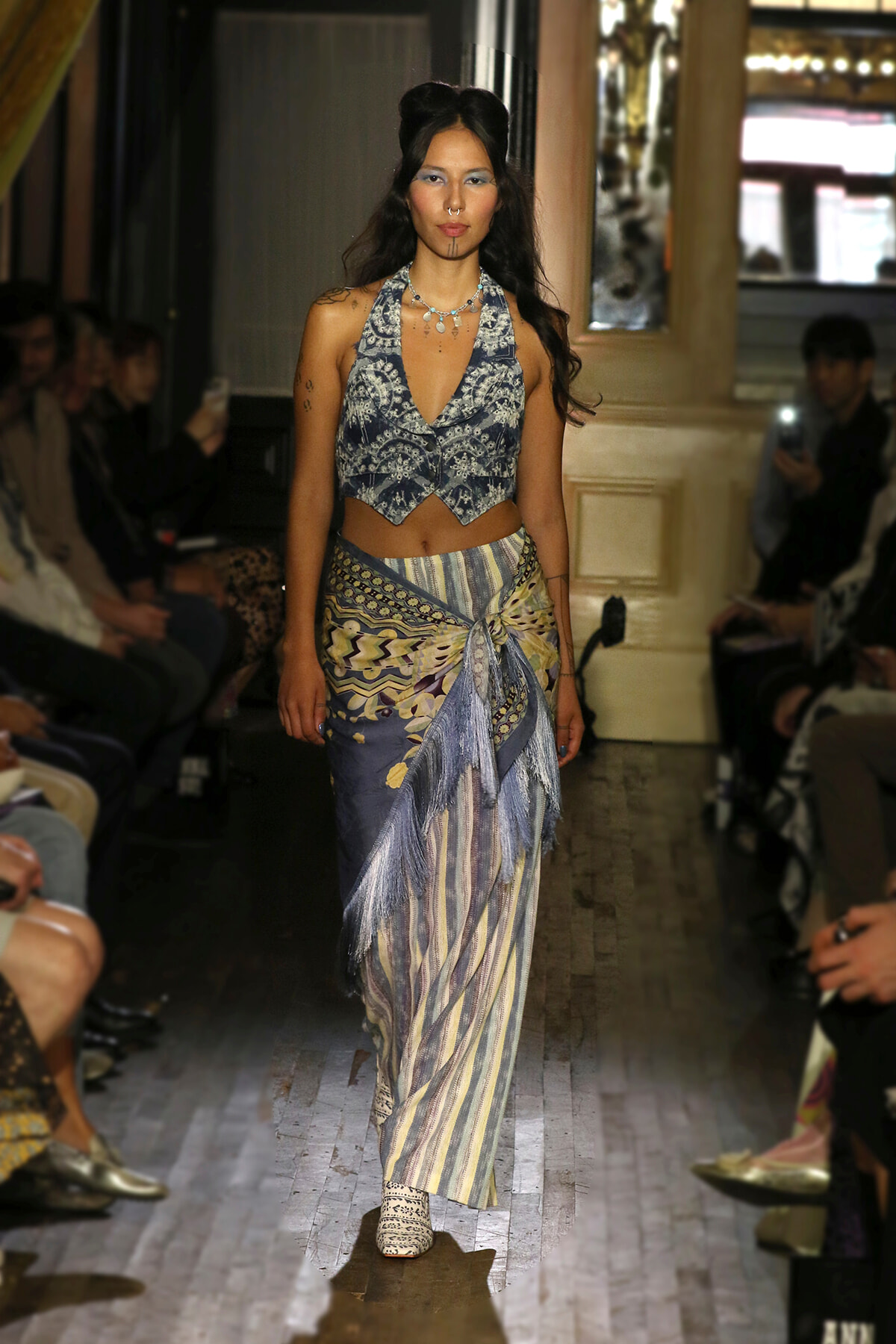 Model walking runway in a patterned halter crop top and layered striped skirt with fringe, in a fashion show setting.