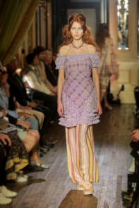 Model walks a fashion runway in a lavender off-the-shoulder lace top with a plaid bodice and striped wide-leg pants; seated audience along the catwalk.
