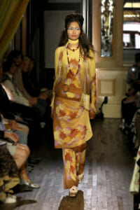 Model walking a fashion runway in a yellow, floral-patterned ensemble with a matching scarf, layered bead necklaces, and a wide brown belt with fringe detailing.