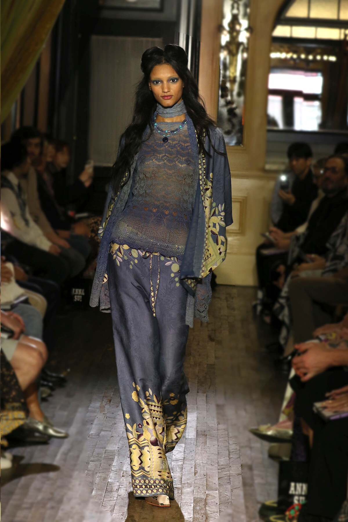 Model walks a runway wearing a blue lace tunic with matching wide-leg pants and a patterned outer layer, accessorized with turquoise beads.