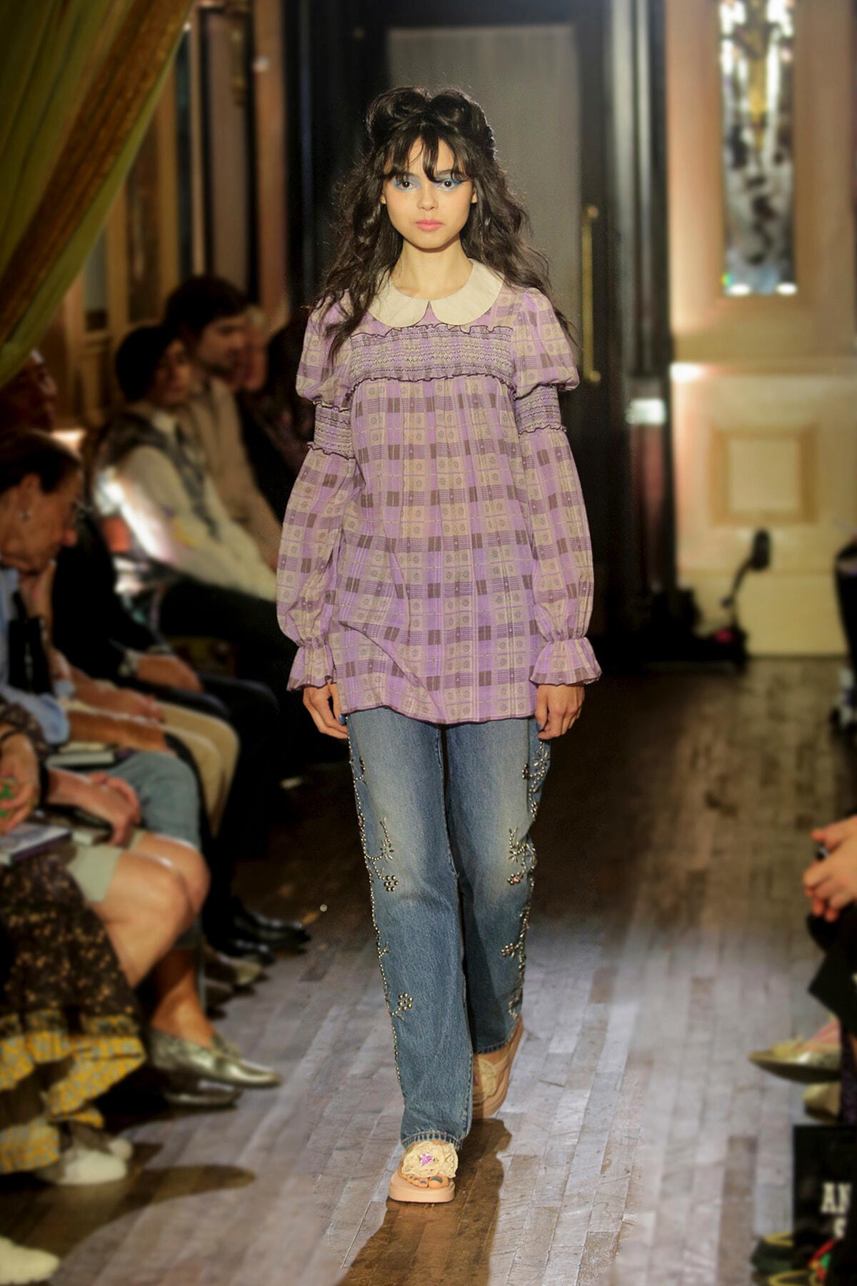 Model on runway wearing a lilac checkered blouse with a white collar and embroidered jeans.