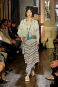 Model walks the runway in a bohemian-inspired outfit: embroidered cream blouse, striped fringe skirt, turquoise accessories, and white sandals.