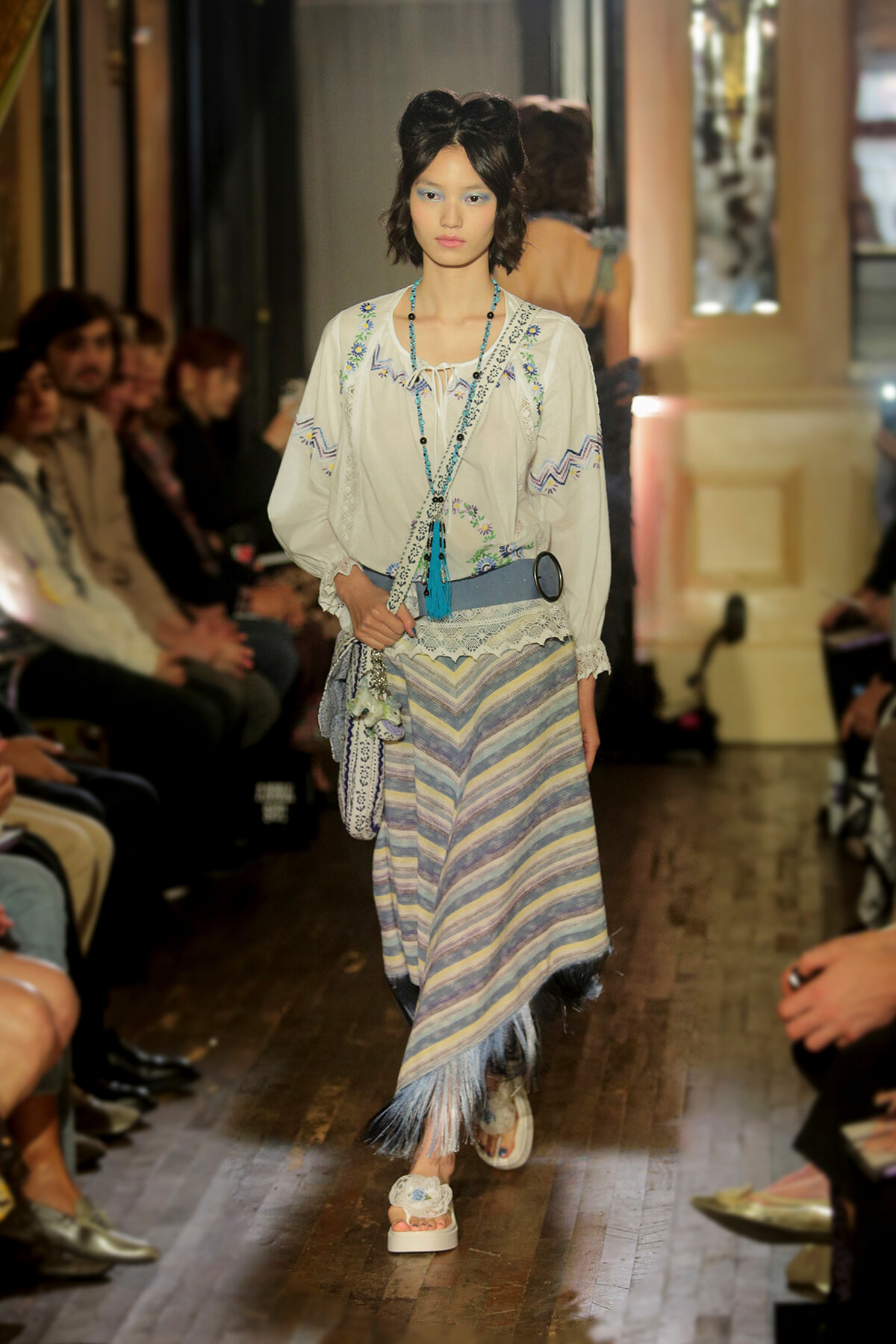 Model walks the runway in a bohemian-inspired outfit: embroidered cream blouse, striped fringe skirt, turquoise accessories, and white sandals.