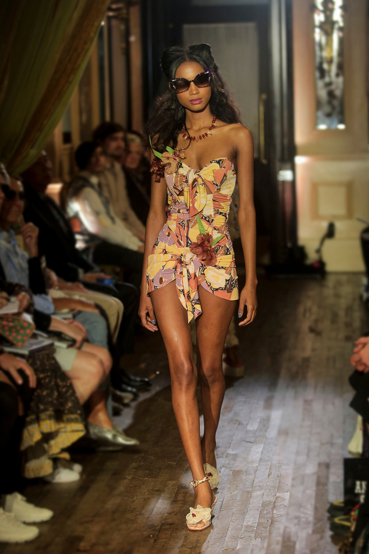Model walking a fashion runway in a floral strapless romper with a large bow, sunglasses, and heels amid a seated audience.