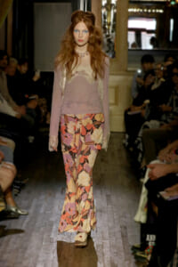 Model walks the runway in a pale lavender lace top with sheer sleeves and a bold floral-print skirt, audience on both sides.