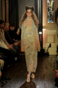 Model walks a runway in a pastel floral, sheer boho dress with scarf, two-bun hairstyle, and pink sunglasses; audience seated along the aisle.