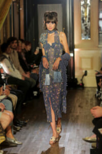 Model walking a runway in a sheer blue floral dress with fringe shawl and floral sandals.