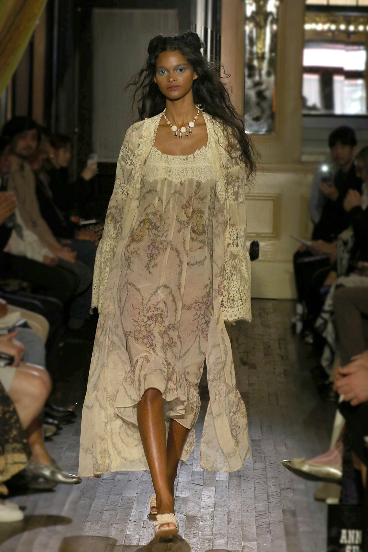 Model walks a runway in a flowing cream lace dress with floral patterns and a chunky shell necklace.