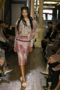 Model walks a runway wearing a pink lace two-piece set—a sheer top with a cropped camisole and matching capri pants—while spectators watch from the sides.