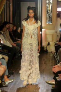 Model walks the runway in a sheer, cream lace gown with floral appliqués and layered pearl necklaces.