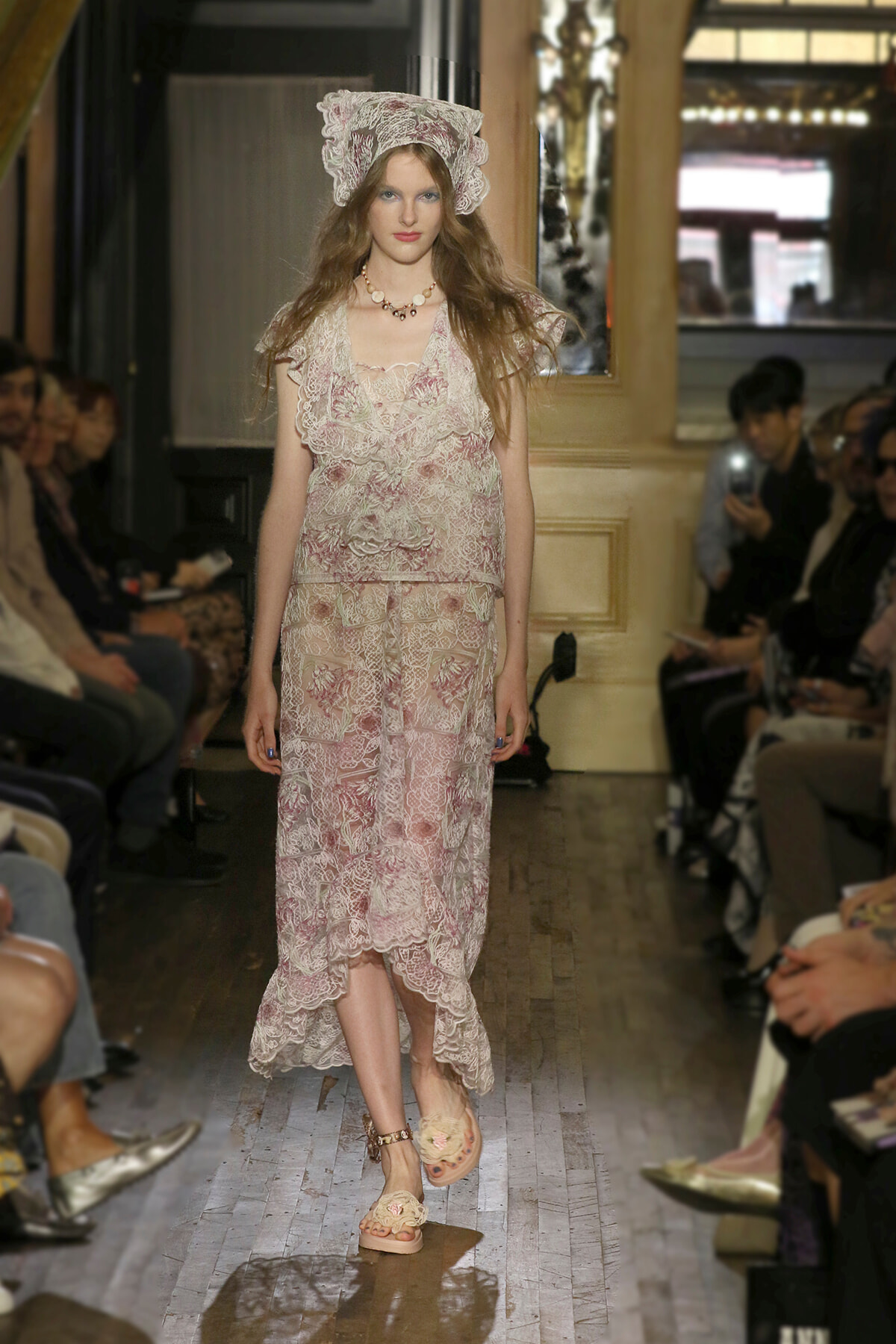 Model walks a runway in a cream-pink lace dress with a matching lace bonnet and beige floral sandals, audience seated on sides behind her.