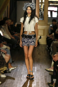 Model walks the runway in a white cropped blouse, blue lace-pattern shorts, and chunky belt with hoop buckle, paired with black sandals and a blue headwrap accessory.