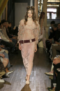 Model walks the runway in a pale pink lace dress with sheer sleeves and a wide burgundy belt, floral details visible throughout the gown (fashion show).