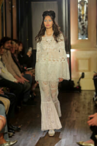 Model walks the runway in a white lace, bohemian-style dress with tiered, sheer layers and floral embroidery.