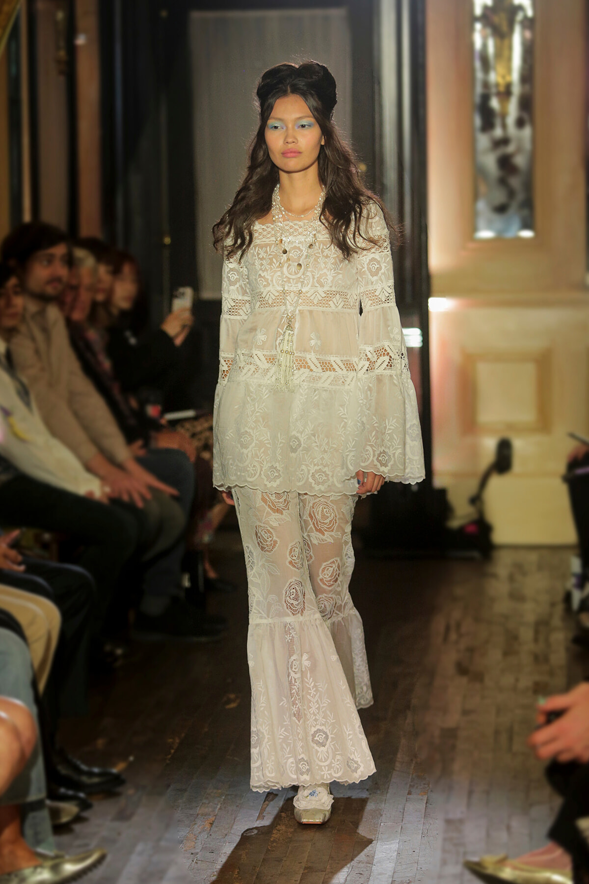 Model walks the runway in a white lace, bohemian-style dress with tiered, sheer layers and floral embroidery.