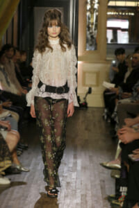 Model walking a runway in a white lace blouse and sheer embroidered black pants with a wide belt.