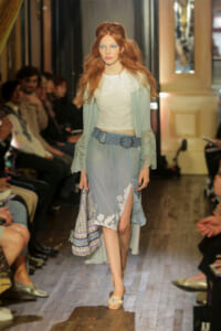 Model walking a runway in a pale blue embroidered skirt, white top, and lace cardigan with a wide belt; audience along the sides.