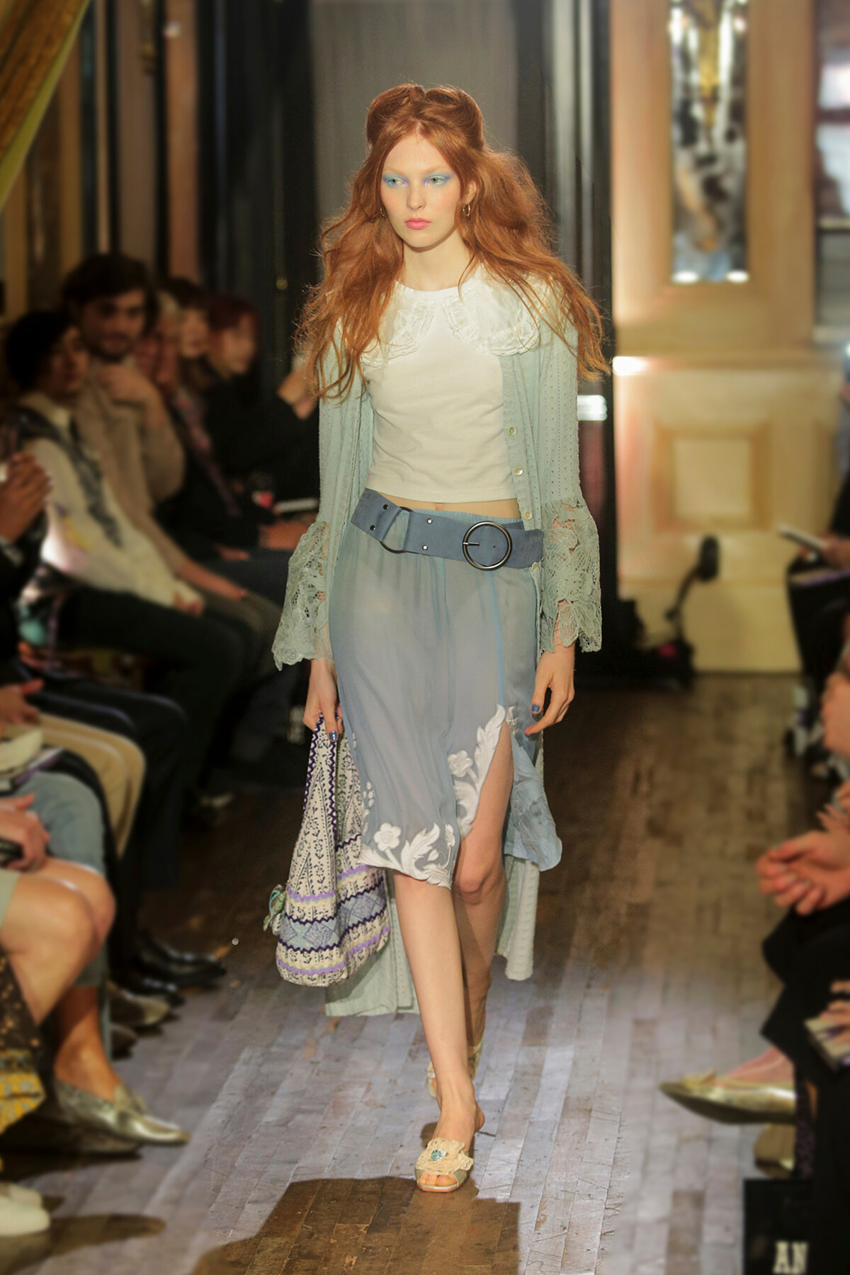 Model walking a runway in a pale blue embroidered skirt, white top, and lace cardigan with a wide belt; audience along the sides.