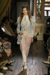 Model walks a fashion runway in a pale blue embroidered jacket and sheer lace pants, accessorized with layered necklaces and a small beaded bag. Audience sits along the sides.