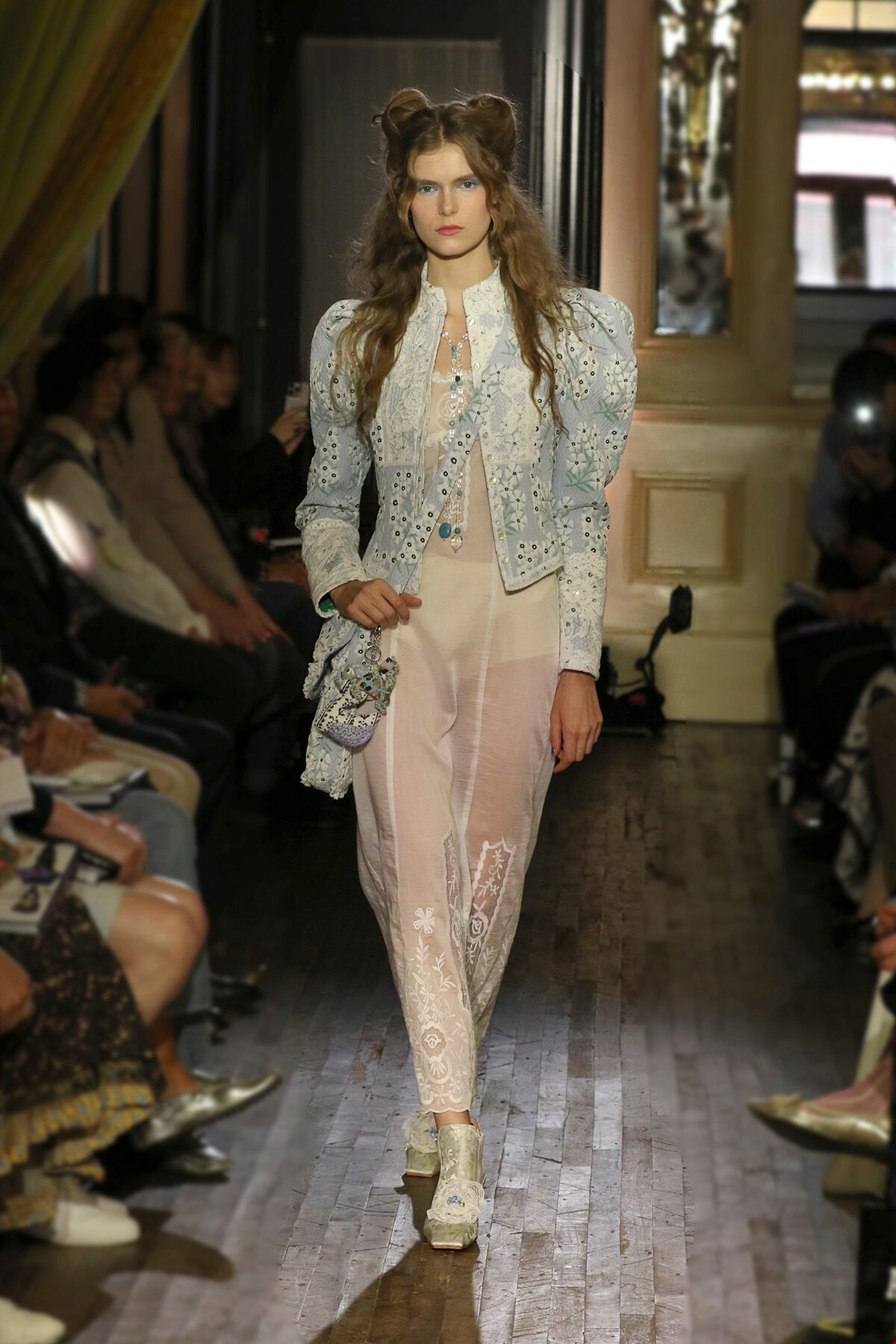 Model walks a fashion runway in a pale blue embroidered jacket and sheer lace pants, accessorized with layered necklaces and a small beaded bag. Audience sits along the sides.