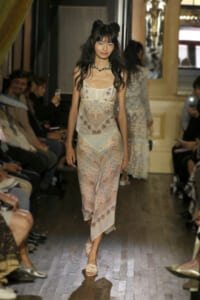 Model walking a runway in a pale patterned dress with a blue beaded necklace, audience on both sides.