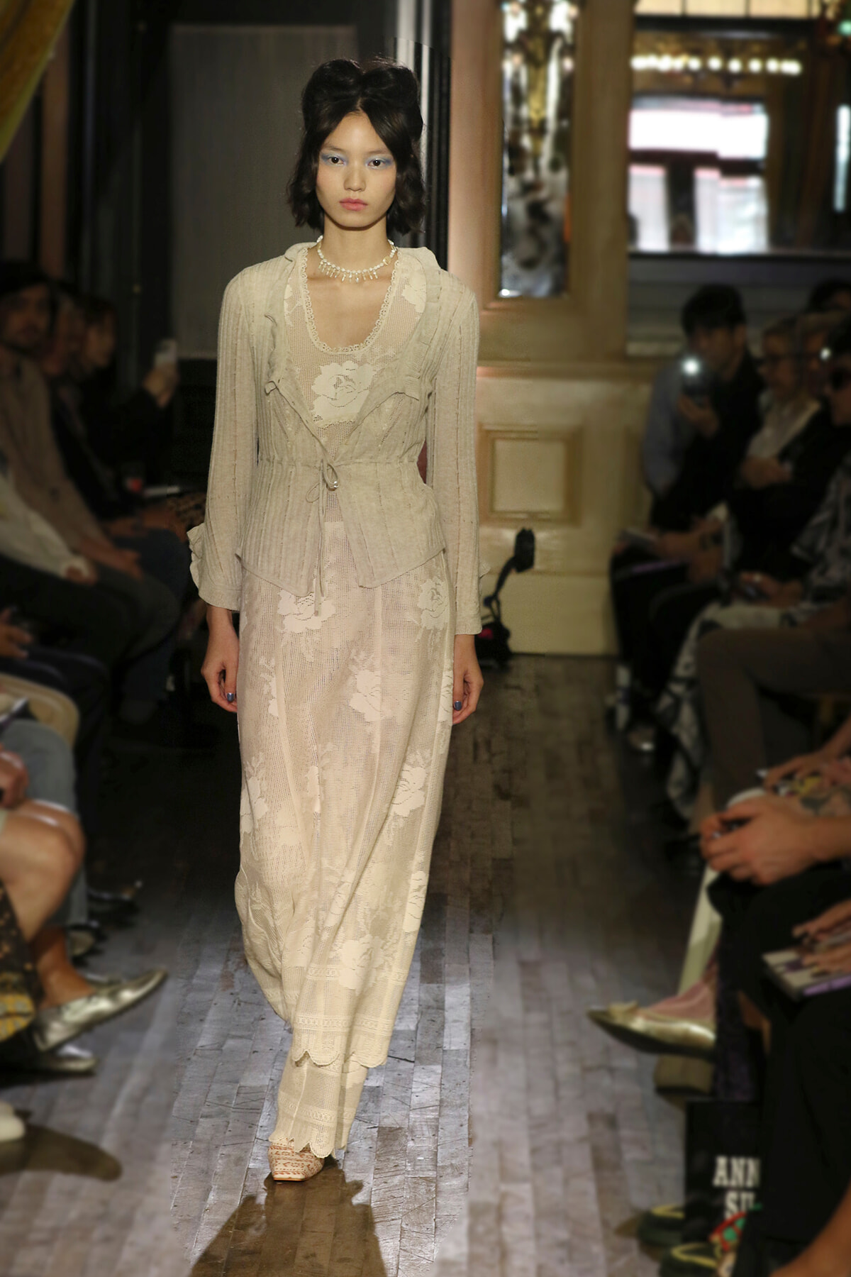 Runway model wearing a cream lace two-piece outfit with a belt, pearls, and sheer floral skirt, walking down a fashion show aisle.