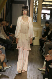 Model walking a runway in pastel pink striped ensemble with ruffled hem, layered necklaces, and sheer bodice top; audience silhouettes in background.