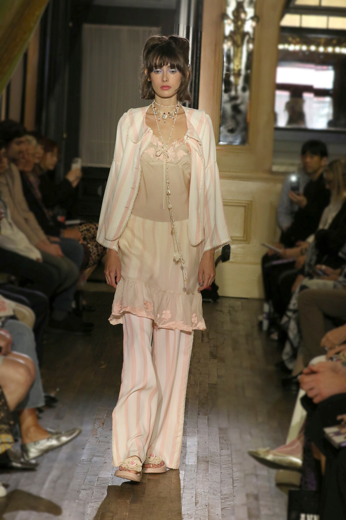 Model walking a runway in pastel pink striped ensemble with ruffled hem, layered necklaces, and sheer bodice top; audience silhouettes in background.